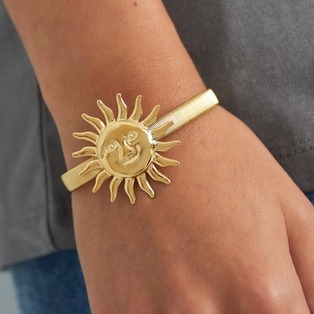Gold sun-shaped bracelet on a wrist with a gray shirt and blue jeans Stainless steel (verkleurd niet) background bynooni bangle