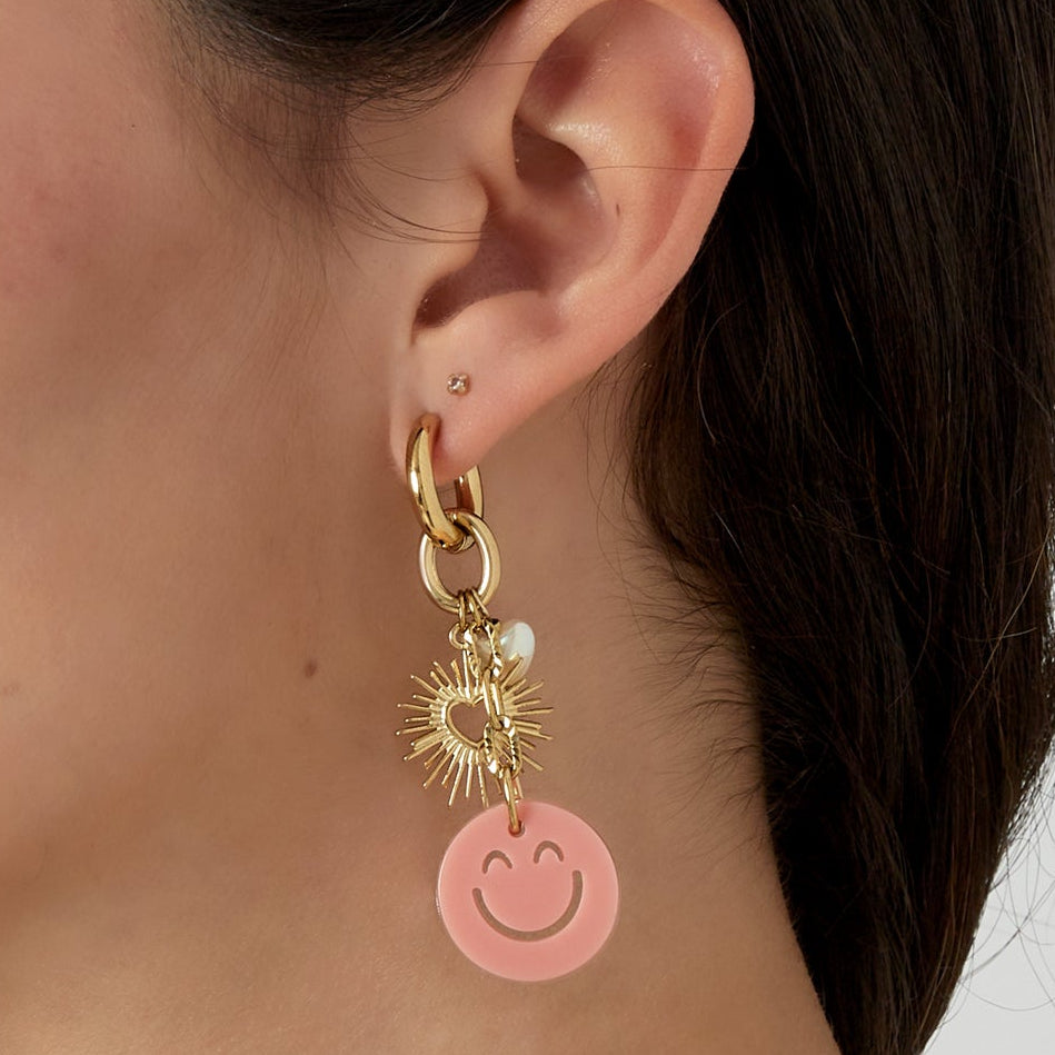 Close-up of a person wearing a gold earring with a pink smiley face charm on a neutral background