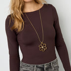 Woman wearing a long-sleeve brown top and gray jeans with a gold necklace featuring a flower pendant on a white background.