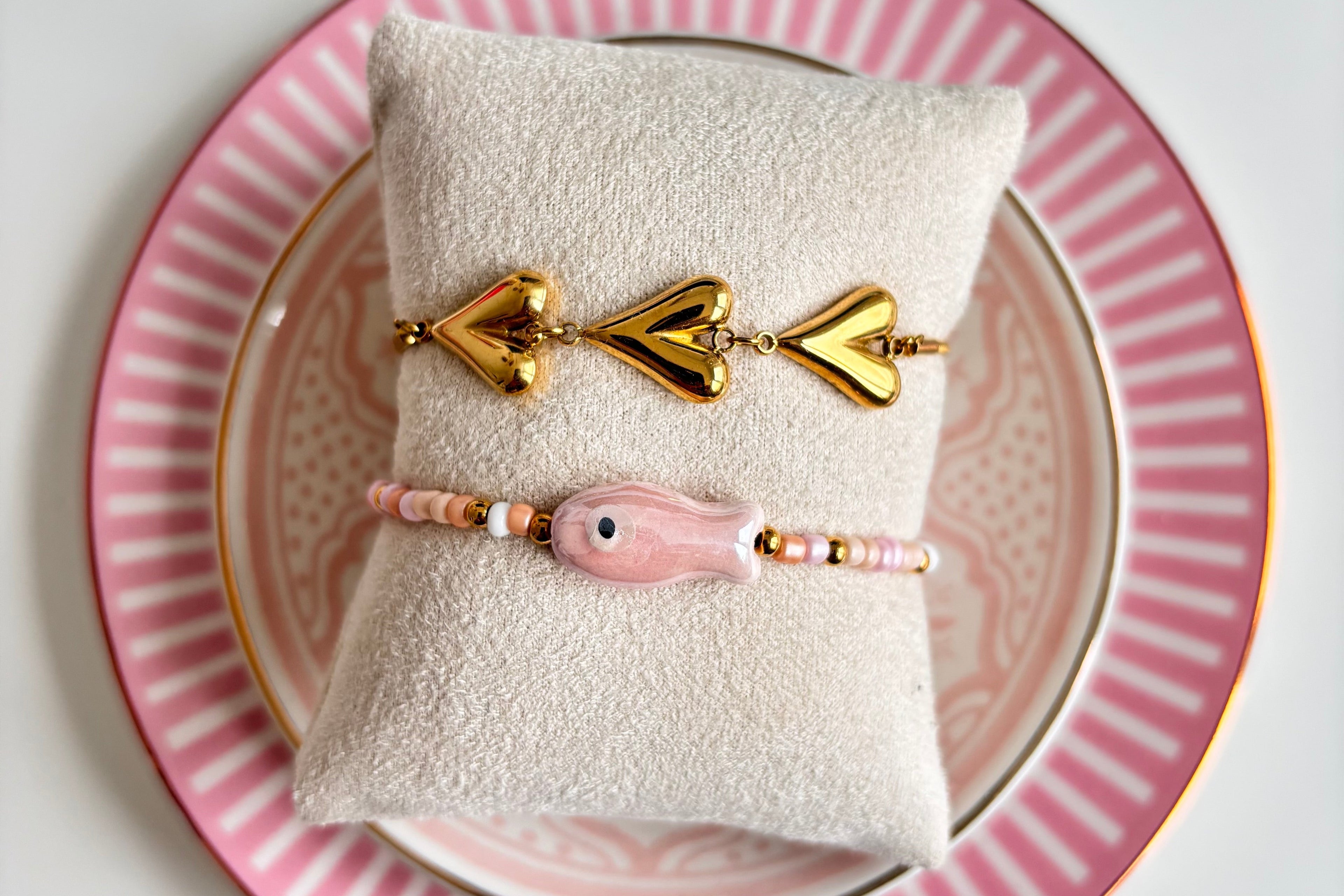 Two gold bracelets on a white pillow with a pink and gold decorative plate underneath.