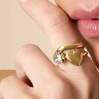 Close-up of a hand wearing a gold ring with a heart-shaped pendant, touching the chin.