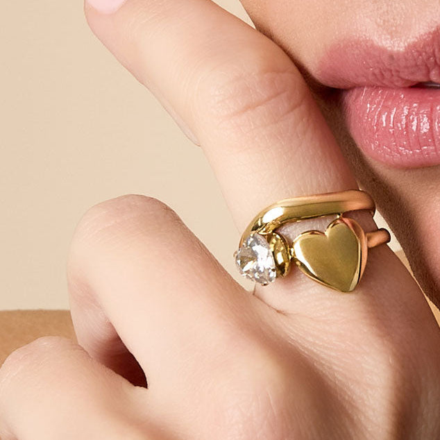Close-up of a hand wearing a gold ring with a heart-shaped pendant, touching the chin.